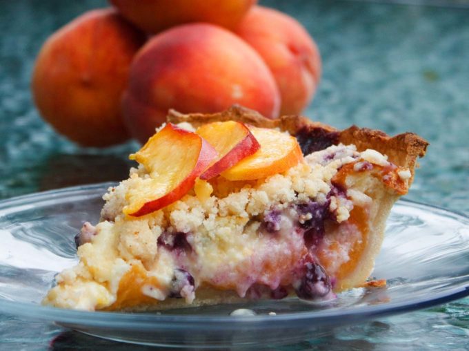 slice of peach blueberry custard pie on a glass plate with a stack of peaches behind it