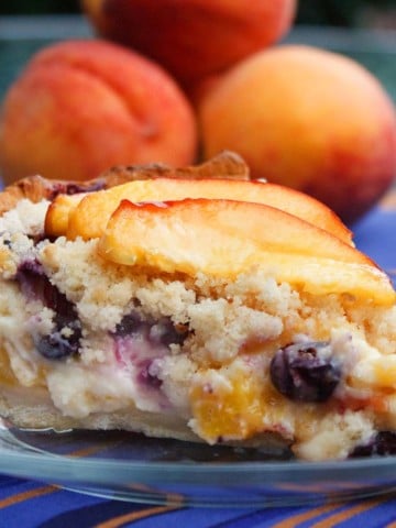 slice of peach blueberry custard pie on a glass plate with a stack of peaches behind it