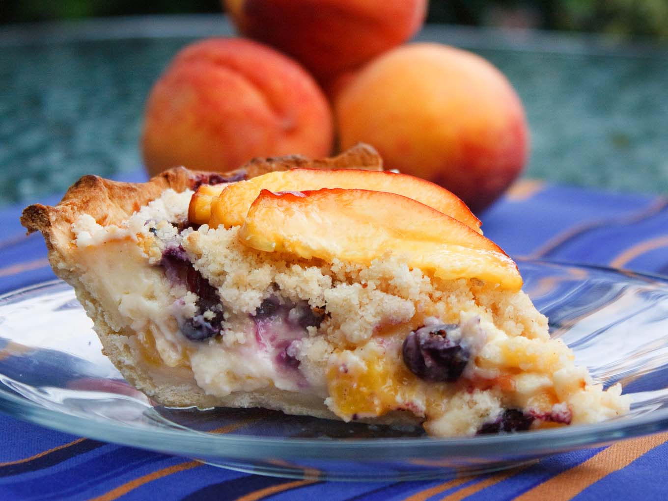 slice of peach blueberry custard pie on a glass plate with a stack of peaches behind it