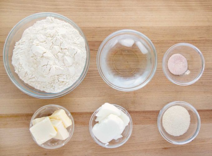 ingredients to make a pie crust in glass bowls sitting on a wooden cutting board