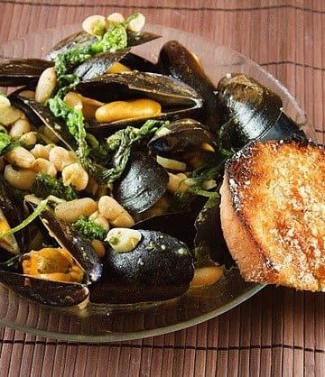 mussels with broccoli rabe and cannellini beans in a large glass bowl with a slice of garlic bread against the bowl