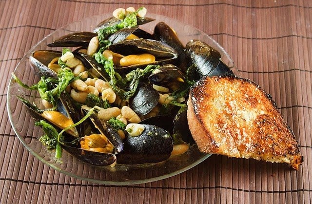 mussels with broccoli rabe and cannellini beans in a large glass bowl with a slice of garlic bread against the bowl