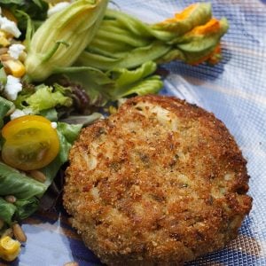 one crab cake next to a salad with zucchini blossoms on a clear plate