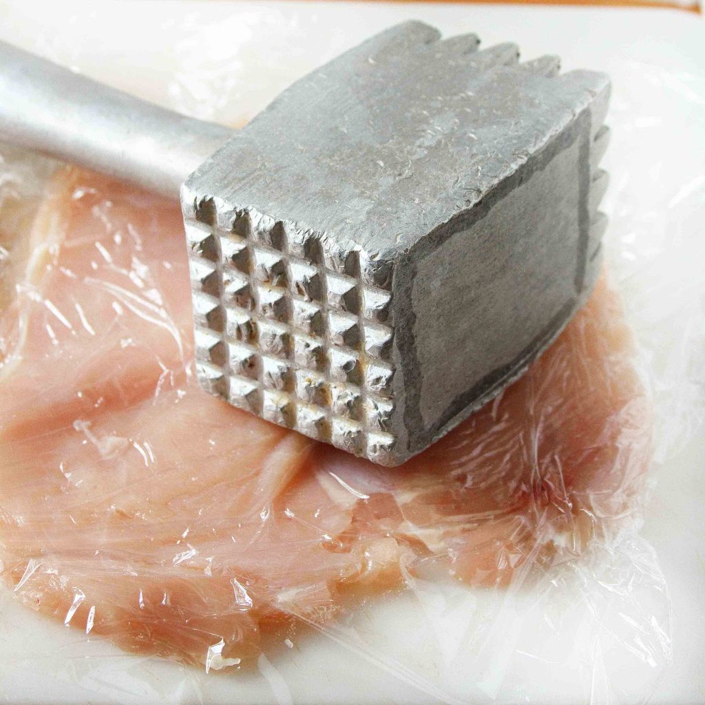 chicken breast between plastic wrap on a cutting board with a meat hammer sitting on top