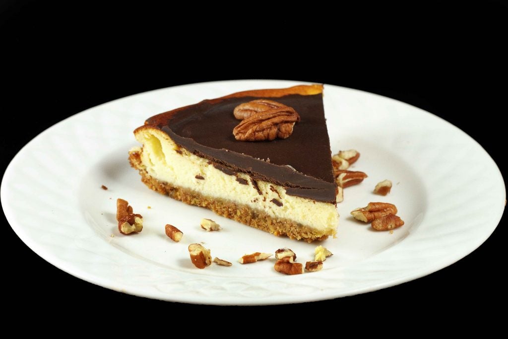 slice of Feta Cheesecake topped with chocolate ganache served on a white plate with crumbled pecans