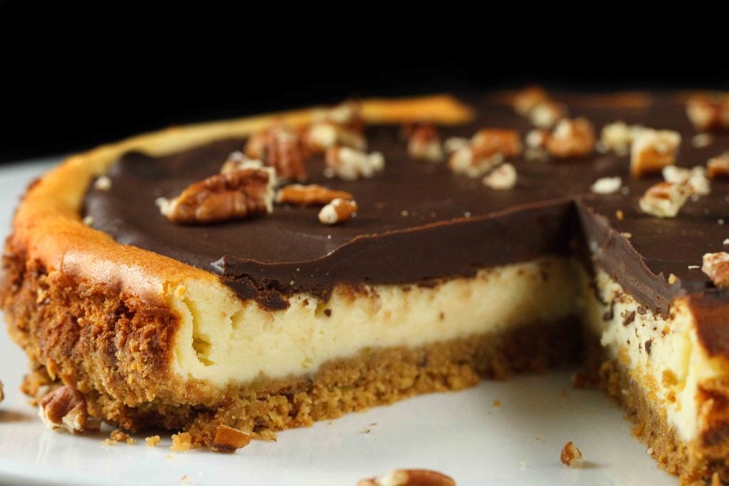 chcocolate topped Feta Cheesecake topped with pecans with a graham cracker crust sitting on a white plate