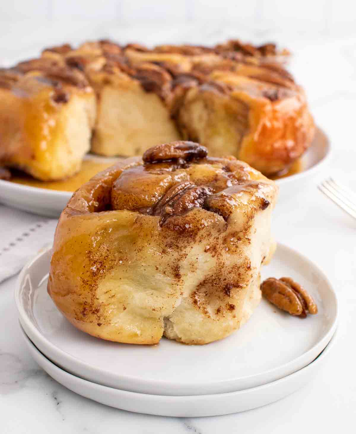 Pecan Caramel Roll 1 Caramel Pecan bun on white plate with platter of buns behind it