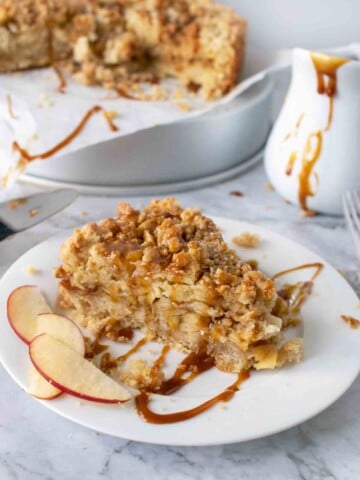 slice of caramel apple torte on a white plate with a white pitcher of caramel and the rest of the cake behind it