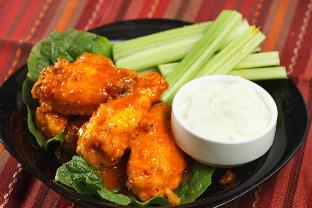 honey lime buffalo wings with blue cheese dressing and celery sticks on a black plate on a multi colored table cloth