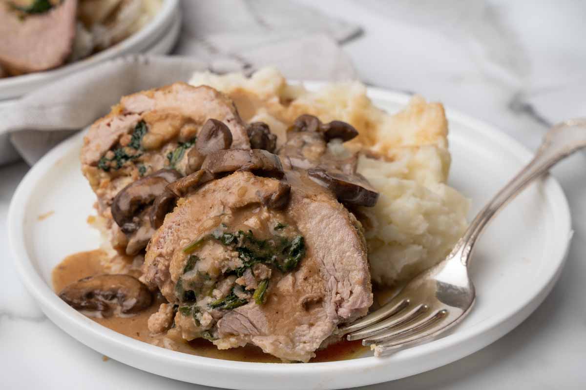 slices of stuffed pork tenderloin on a white plate with mushroom sauce and mashed potatoes