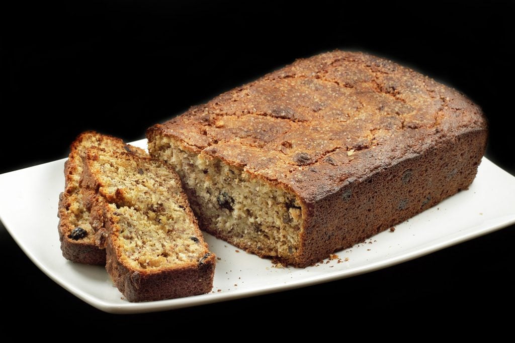 Blueberry oat Banana bread with Pecans with two slices sitting in front of the whole cake on a white plate on a black background