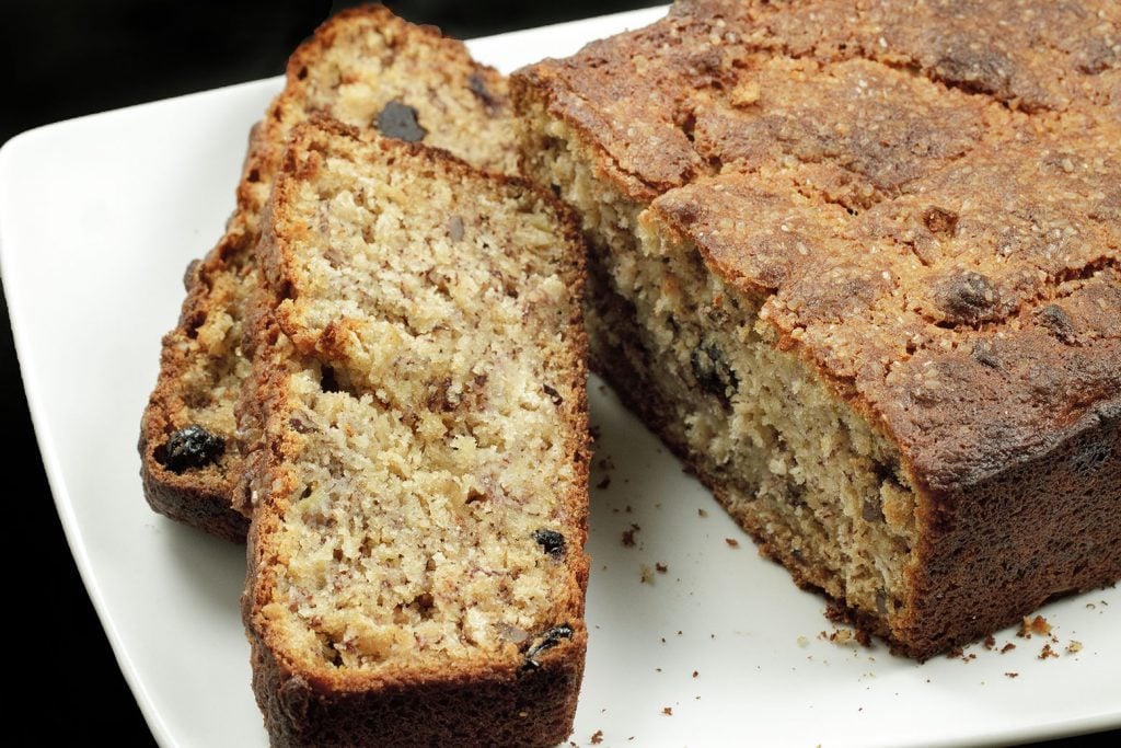 Blueberry oat Banana bread with Pecans with two slices sitting in front of the whole cake on a white plate on a black background