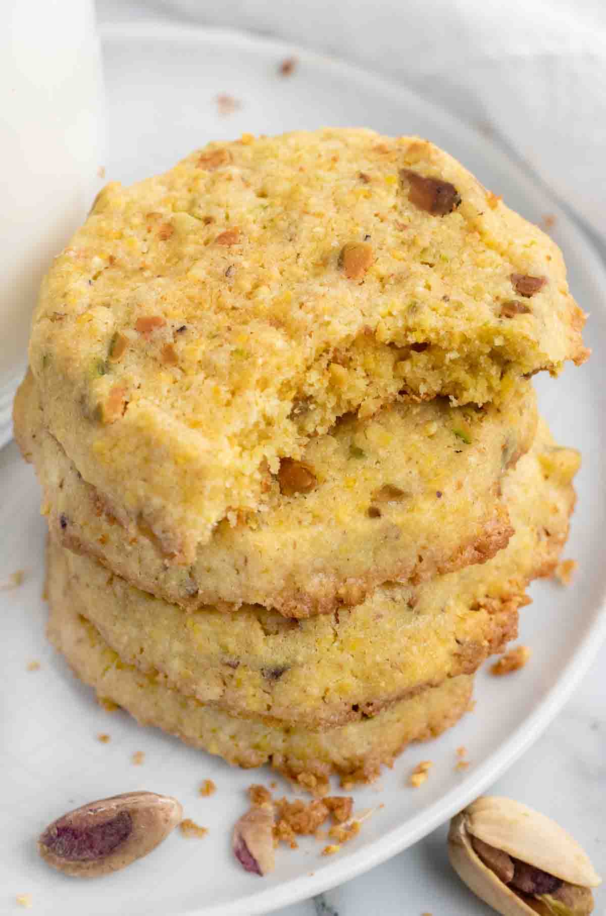 stack of pistachio cornmeal cookies with a bite out of the top one on a white plate