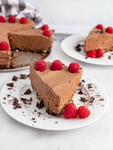 slice of no bake chocolate cheesecake on a white plate with another slice and the whole cheesecake behind it