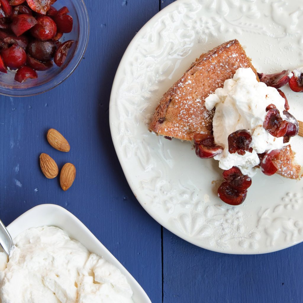 overhead partial view of a slice of cherry almond cake on a white plate with bowls of cherries and whipped cream