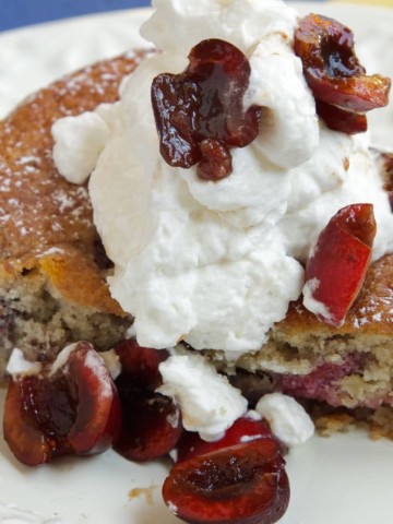 slice of cherry almond cake topped with whipped cream and sliced cherries on a white plate