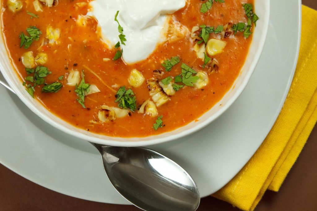 partial view of roasted red pepper soup in a white bowl on a white plate with a spoon