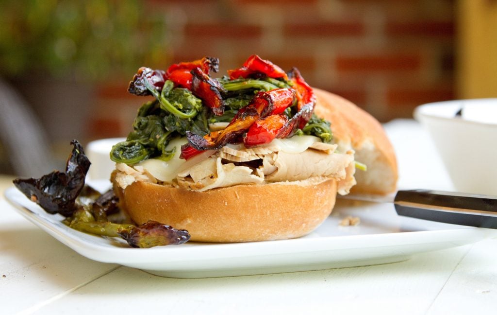 Philly style roast pork side view of roast pork sandwich with provolone cheese, roasted red peppers and broccoli rabe sitting on a white plate on a white table with a steak knife on the plate