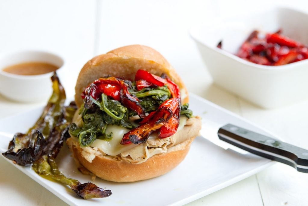  roast pork sandwich with provolone cheese, roasted red peppers and broccoli rabe sitting on a white plate on a white table with a steak knife on the plate and a bowl of roasted red peppers and ramekin of au jus in the background