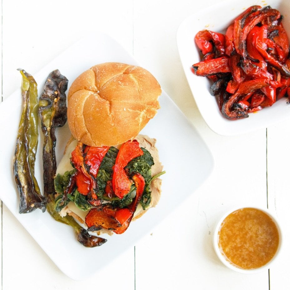 overhead view of roast pork sandwich with provolone cheese, roasted red peppers and broccoli rabe sitting on a white plate on a white table with a bowl of roasted red peppers and small ramekin of au jus