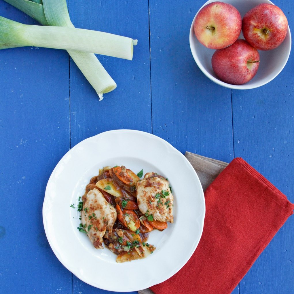 Jack Daniels apple butter chicken on a white plate with a Red Napkin underneath, a bowl of red apples and 2 leeks crossed all sitting on a bright blue table