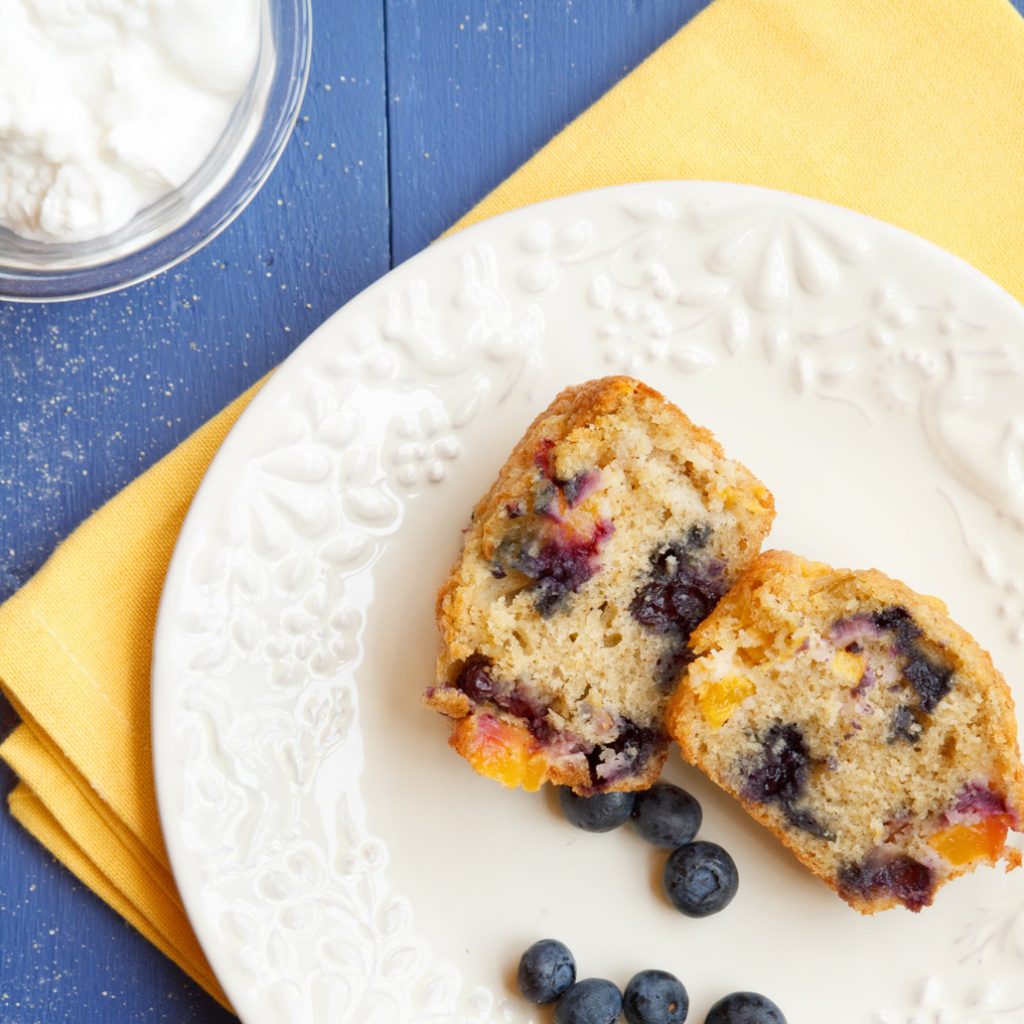 nectarine blueberry muffins sitting on a white plate on a yellow napkin and a glass bowl of whipped cream sitting on a blue table