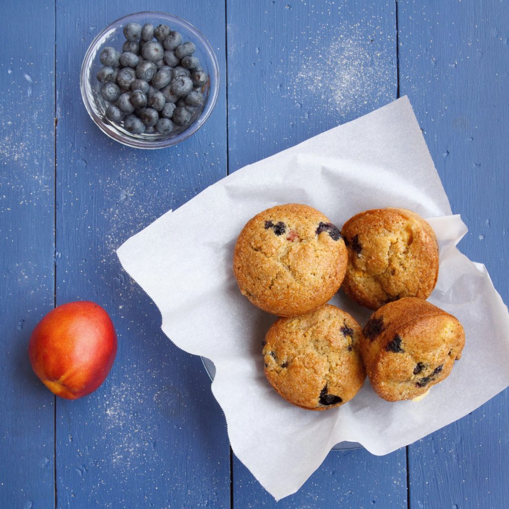 nectarine blueberry muffins sitting on a white paper with a nectarine and glass bowl of blueberries all sitting on a blue table