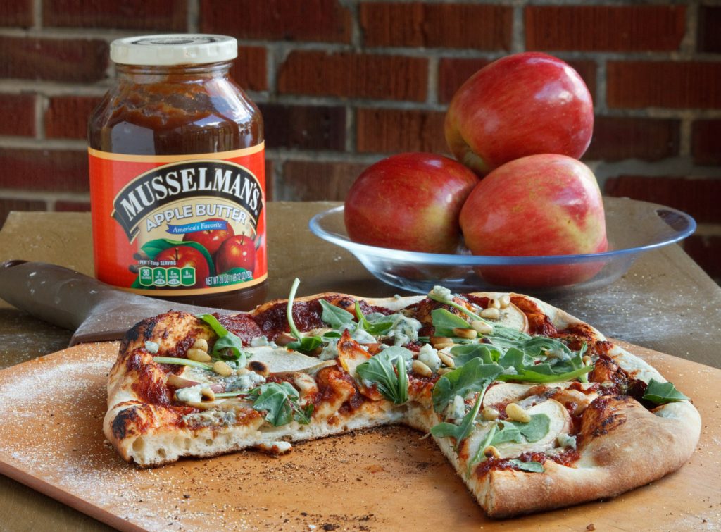 apple butter pizza with a slice cut out sitting on a pizza peel. A jar of apple butter and a bowl of apples behind them.