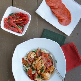 chicken and shrimp pepperoni in a white bowl on a brown table with a dark green napkin and red napkin next to a bowl of red peppers and a plate of sliced pepperoni