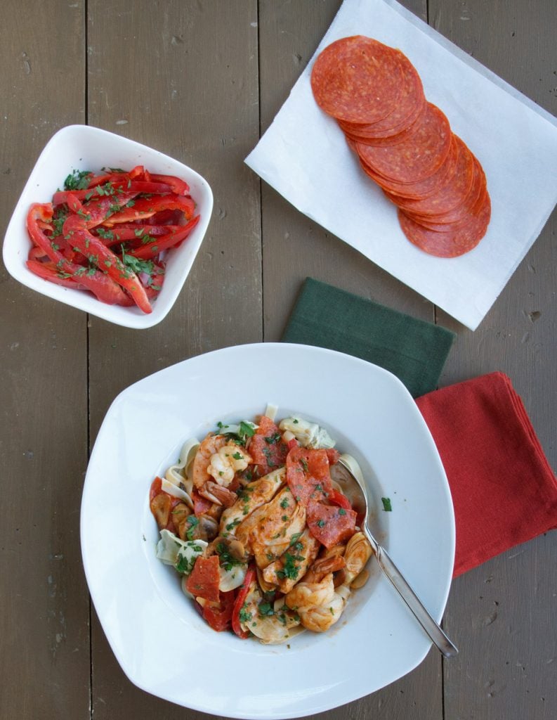 chicken and shrimp pepperoni in a white bowl on a brown table with a dark green napkin and red napkin next to a bowl of red peppers and a plate of sliced pepperoni