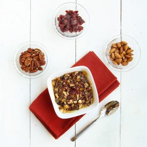 white bowl of dulce de Leche quinoa on top of a red napkin with bowls of fruit and nuts around it