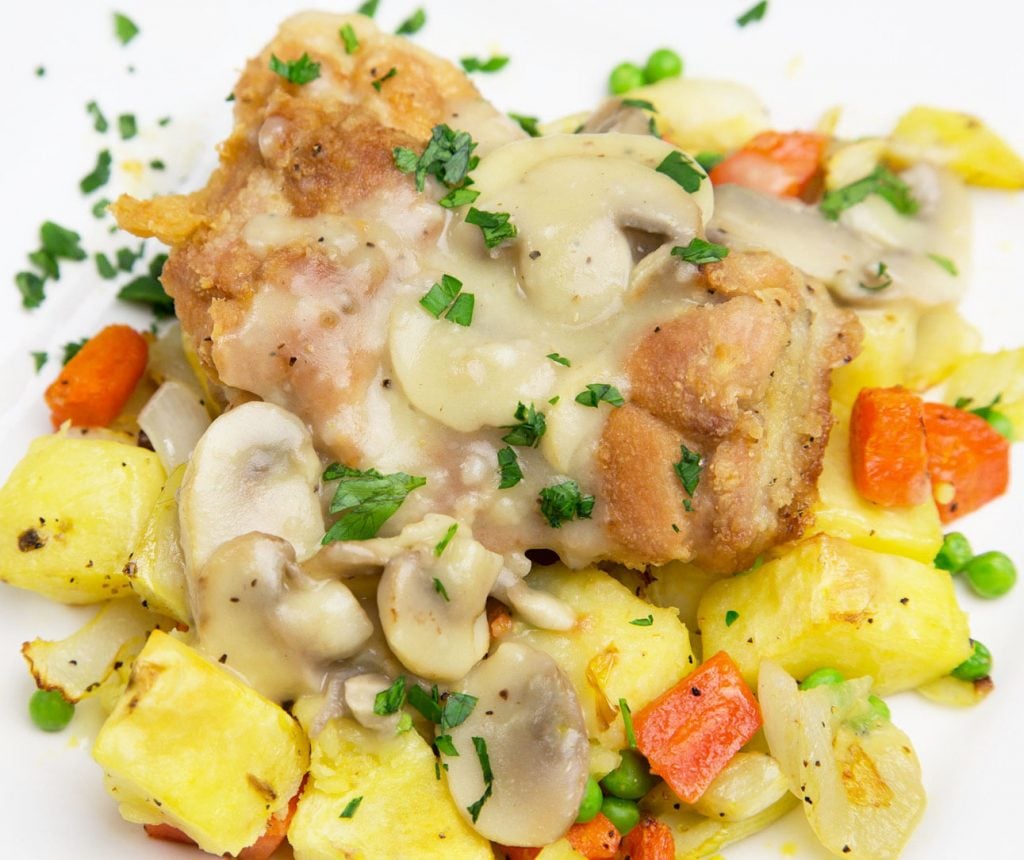 close up of Chicken on a bed of potatoes, carrots, peas and other vegetables topped with mushroom sauce on a white plate 