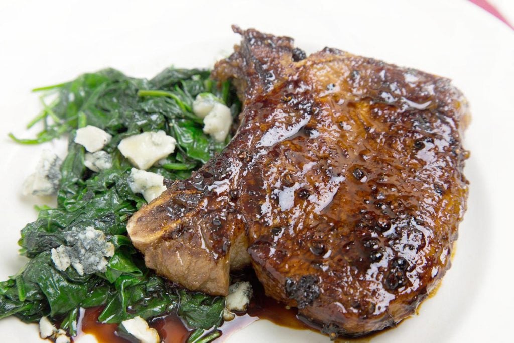 Balsamic Glazed Pork Chop sitting on a white plate with a side of greens with gorgonzola cheese crumbles