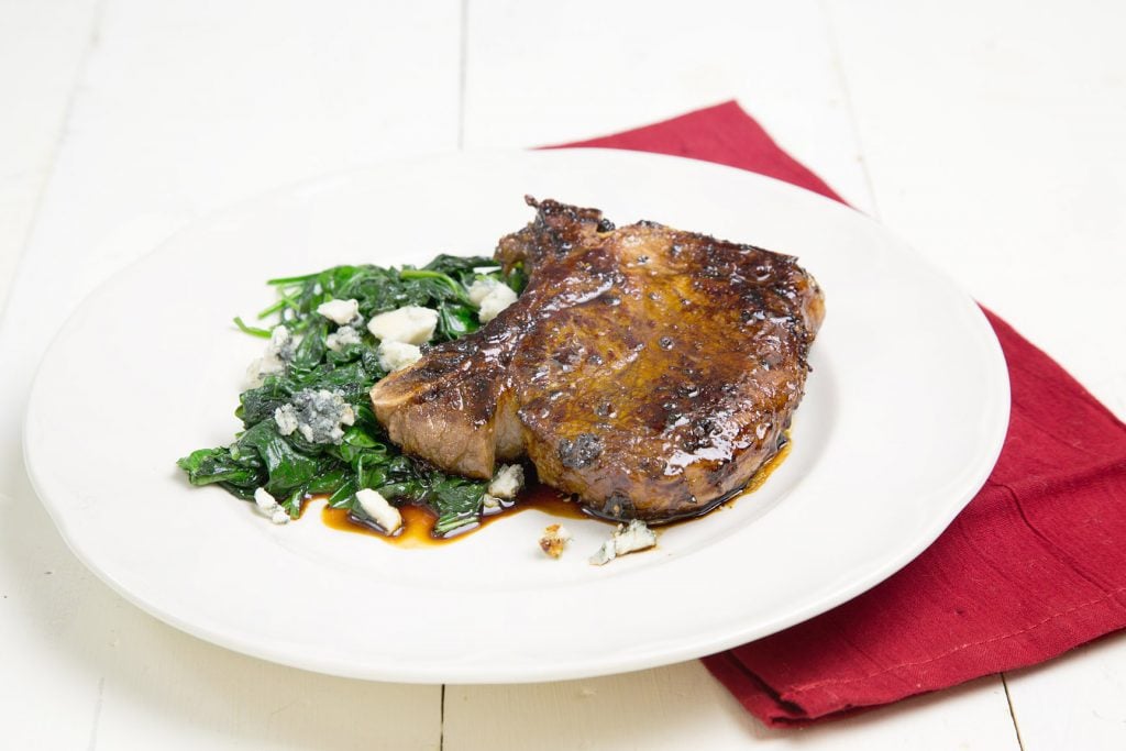 Balsamic Glazed Pork Chop sitting on a white plate on a white table with a side of greens and a red napkin under it