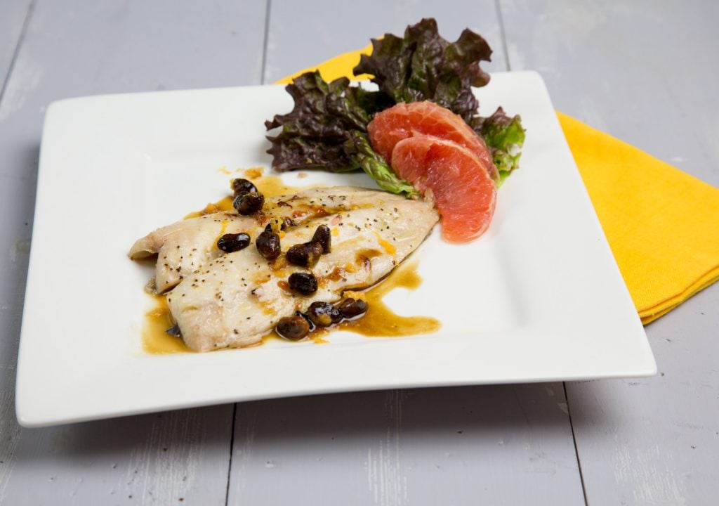 roasted Florida Pompano with pistachios in a sauce on a white plate with sliced grapefruit and red leaf lettuce on the side