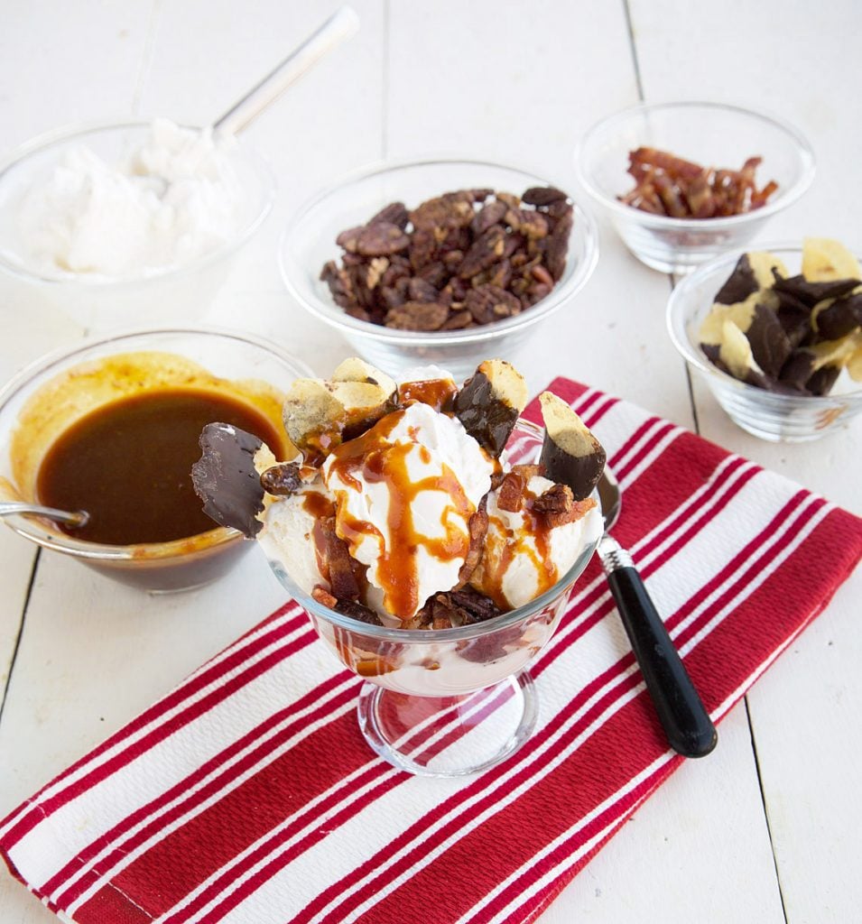 Sundae with Maple Candied Bacon on a red striped napkin, next to bowls of caramel, pecans, choco dipped chips and bacons