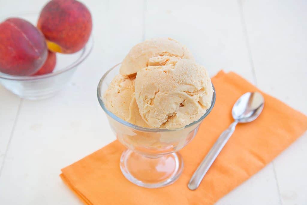 grilled peaches and cream gelato in a glass dish on a tangerine colored napkin with a spoon . A bowl of peaches next to the dish