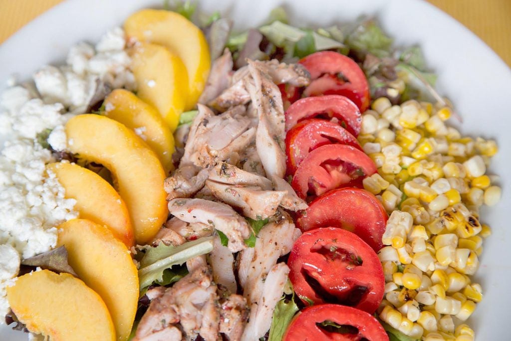 partial overhead view of a salad of feta cheese, sliced peaches, grilled chicken, sliced tomatoes and corn on a white plate