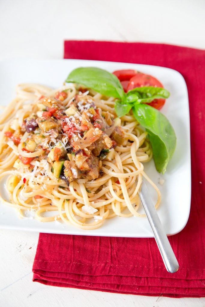 Pasta with vegetables on a white plate with a sprig of basil and a fork, sitting on a red napkin