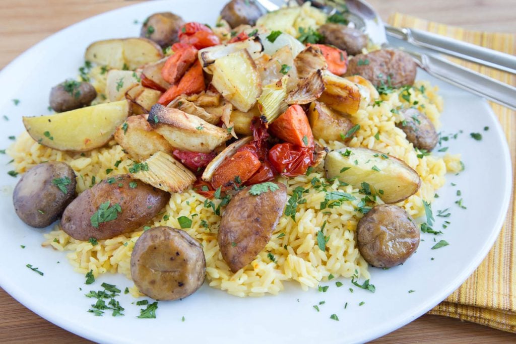 side view of White platter with saffron rice and root vegetables made paella style on a wooden cutting board with a pale yellow striped napkin and a serving spoon and fork on the plate