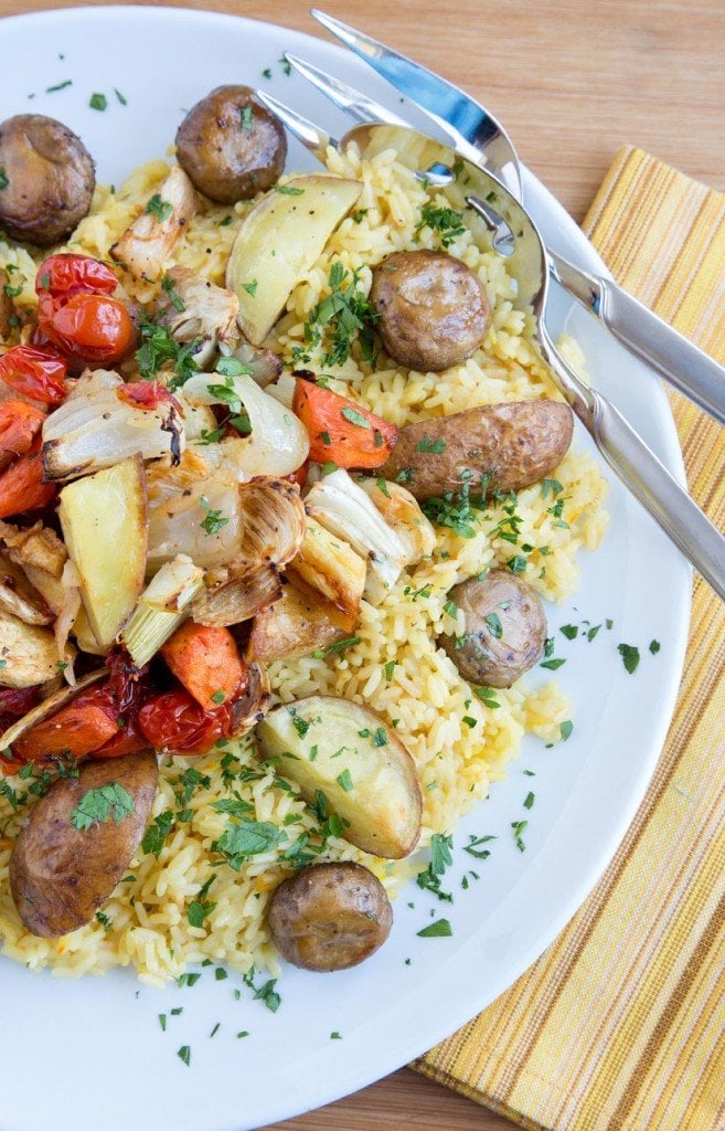 White platter with saffron rice and root vegetables made paella style on a wooden cutting board with a pale yellow striped napkin and a serving spoon and fork on the plate