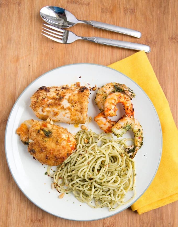 white plate with grilled shrimp, pasta and chicken rollantine with a yellow napkin and silverware sitting on a wooden cutting board