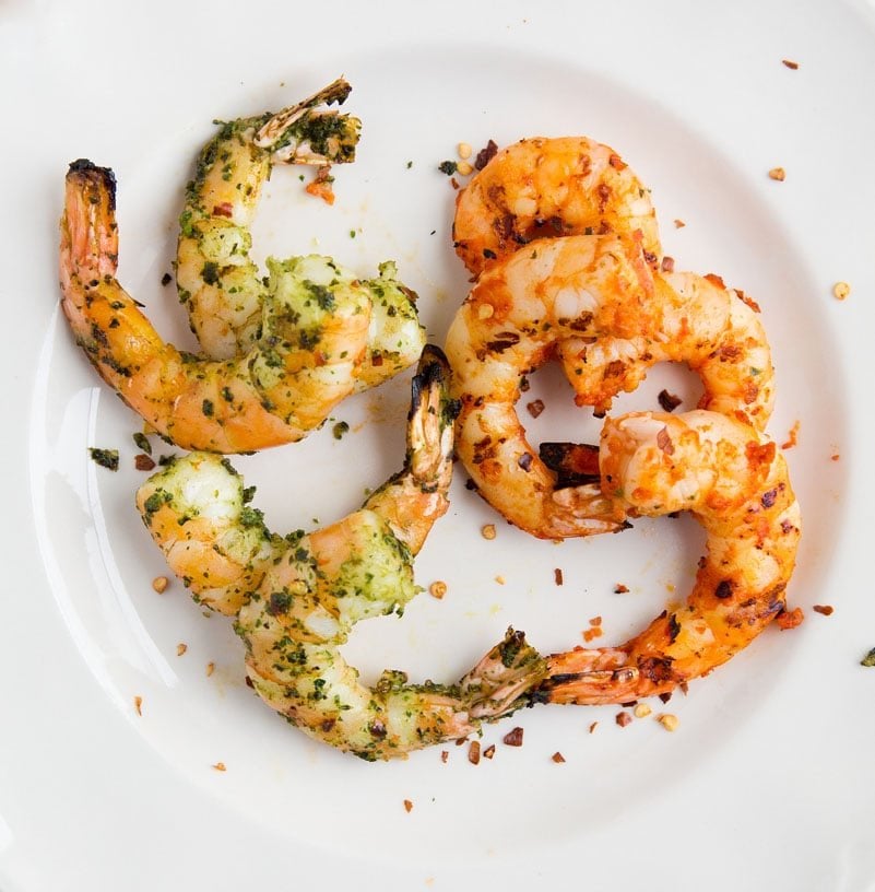 eight grilled shrimp sitting on a white plate. four of them seasoned with basil pesto and four of them seasoned with sundried tomato pesto 