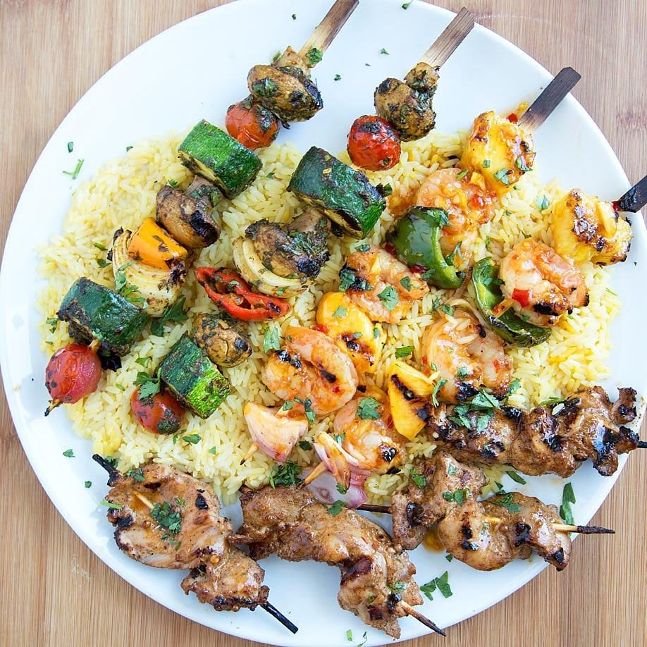 Trio of grilled skewers - Moroccan Vegetable Skewers, Sweet and Spicy Shrimp Skewers and tandoori chicken skewers on a bed of rice on a white plate on top of a cutting board