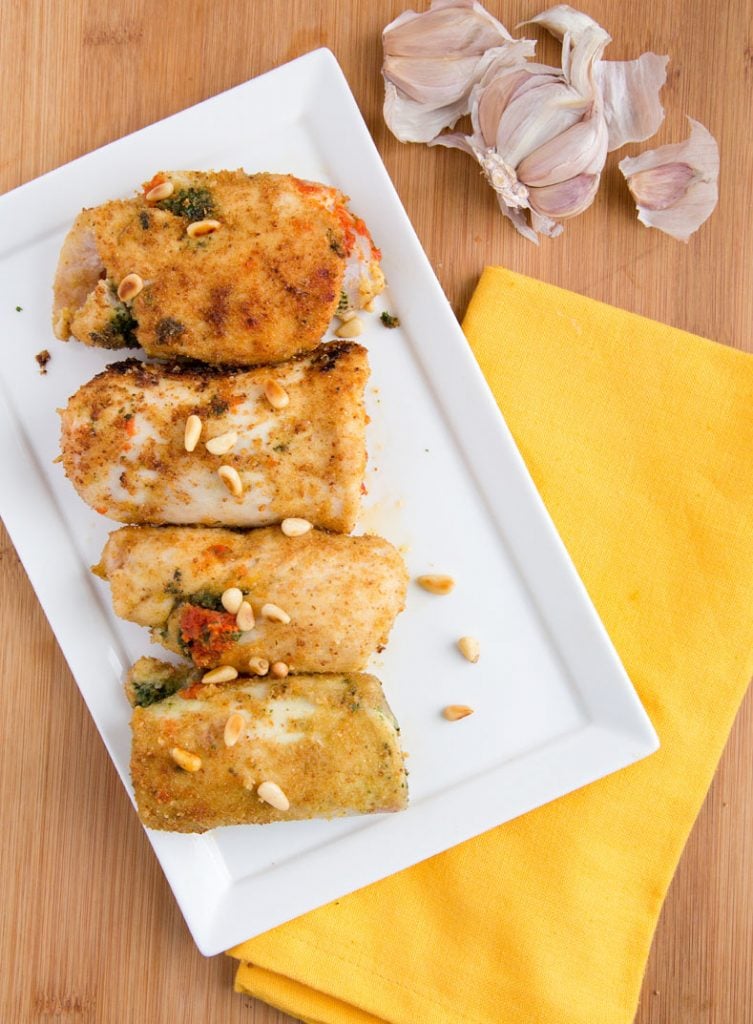 4 sauteed chicken rollantine sitting on a rectangular white plate with toasted pine nuts sprinkled on top sitting on a wooden cutting board with a yellow napkin under a corner of the plate and a head of garlic broken open next to the plate