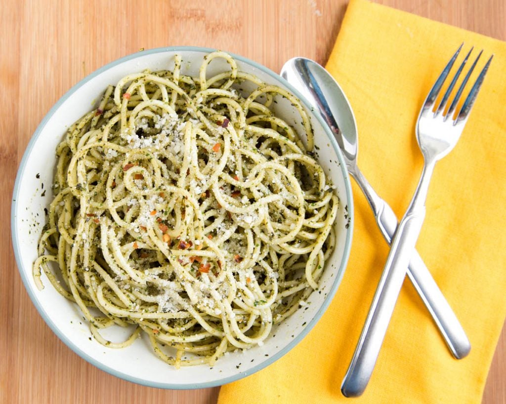 white bowl with a green rim filled with spaghetti alla Pesto Genovese sitting next to a yellow napkin with a spoon and fork crossed on top of the napkin. All sitting on a wooden cutting board