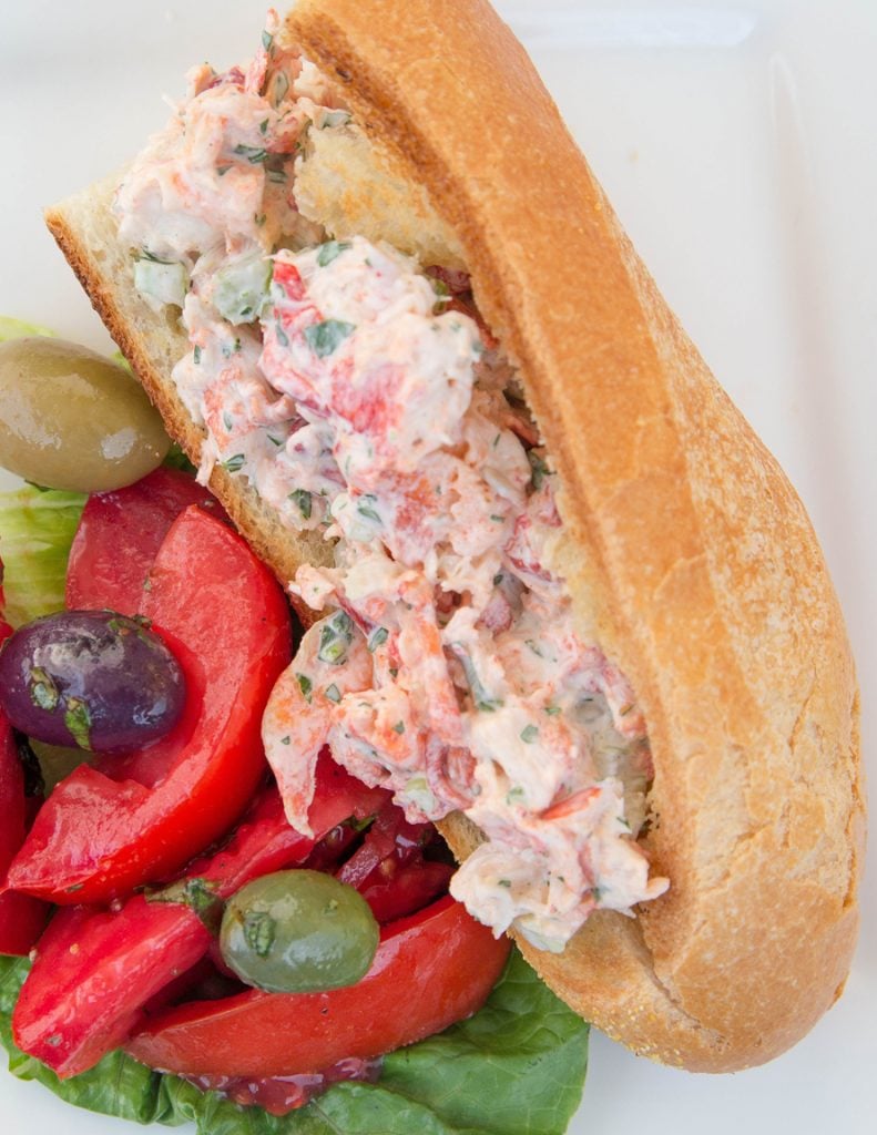 lobster salad on  long roll with a tomato and olive salad on a white plate