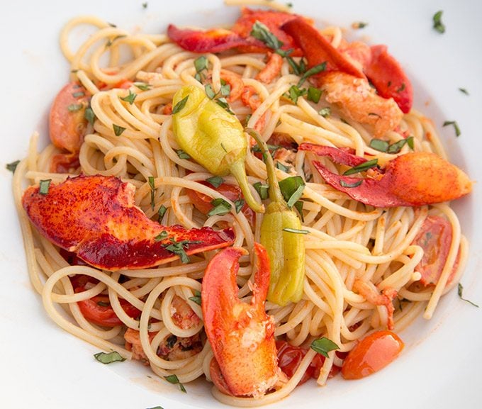 overhead view of lobster claws and lobster meal with pepperoncini over pasta with chopped basil in a white bowl