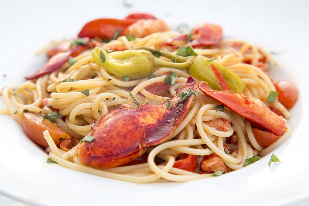 side view of lobster claws and lobster meal with pepperoncini over pasta with chopped basil in a white bowl