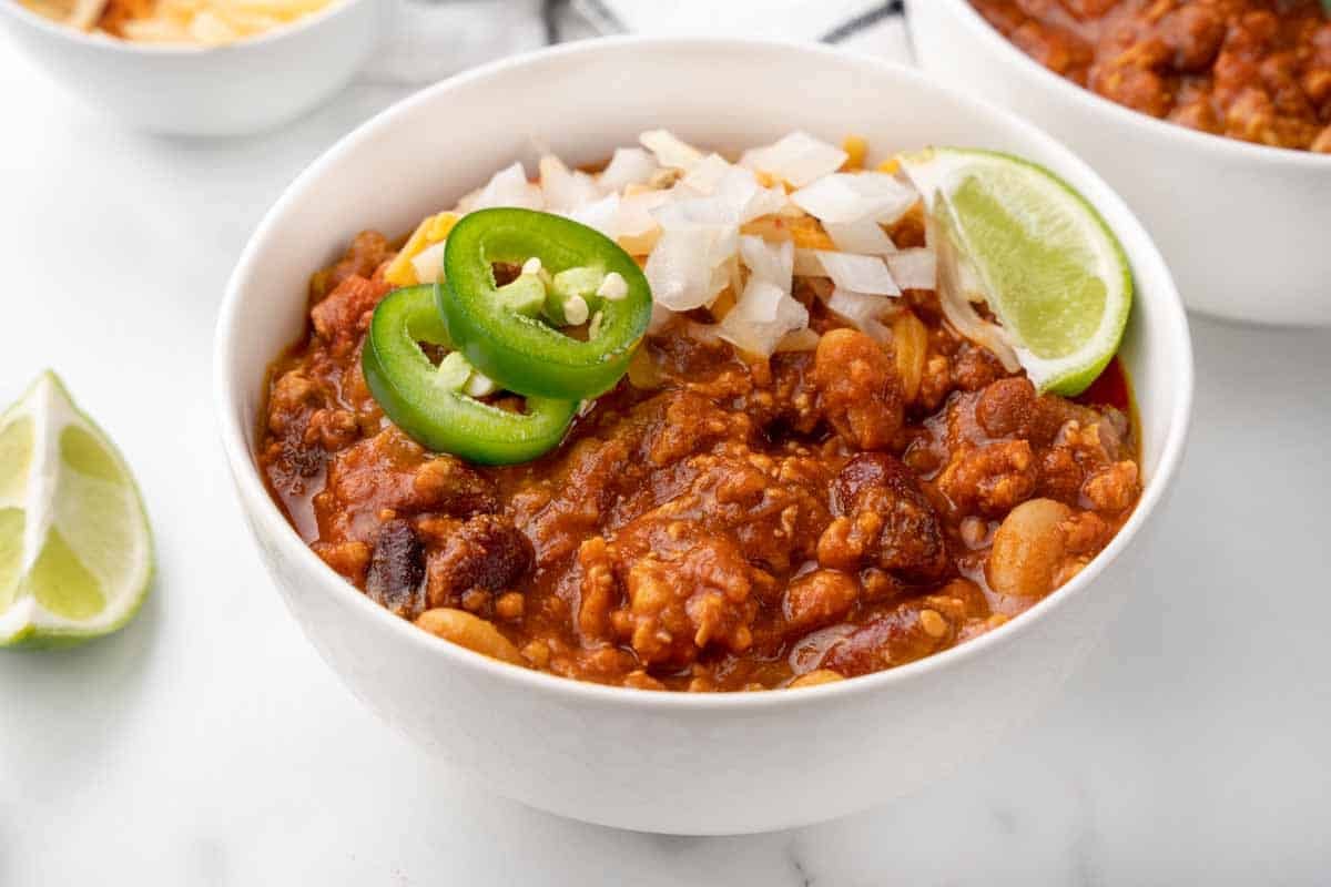 Chicken Chili in a white bowl with shredded cheddar, chopped onion and jalapeno slices